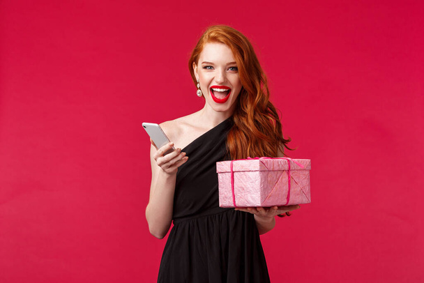 Portrait of lucky and sassy gorgeous redhead woman in red lipstick, black dress, showing happiness and joy, smiling daring look camera, hold gift box and smartphone, red background - Фото, изображение