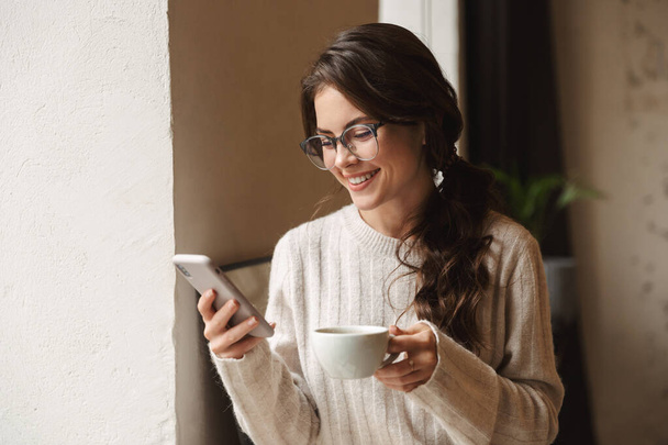 Image of young beautiful caucasian woman with long brown hair holding cellphone and drinking coffee in cafe - Foto, afbeelding