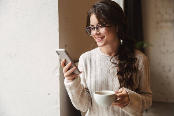 Image of young beautiful caucasian woman with long brown hair holding cellphone and drinking coffee in cafe - Valokuva, kuva