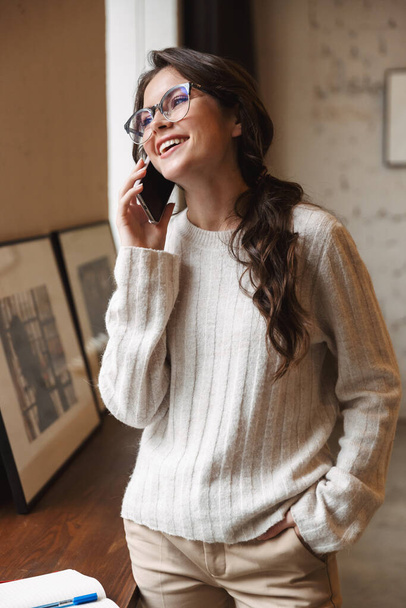 Image of young beautiful caucasian woman wearing eyeglasses talking on smartphone while working in cafe - Foto, Bild