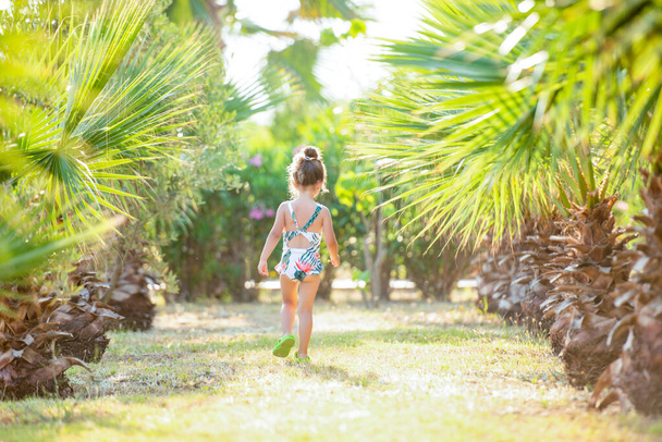 Cute little 3 year old girl in a swimsuit among tropical palm trees. Palm grove. - Zdjęcie, obraz