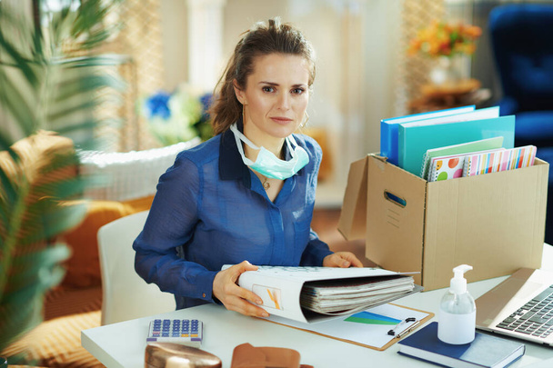 Portrait of stylish female in blue blouse with medical mask organising a temporary home office during the coronavirus epidemic in the modern house in sunny day. - Фото, изображение