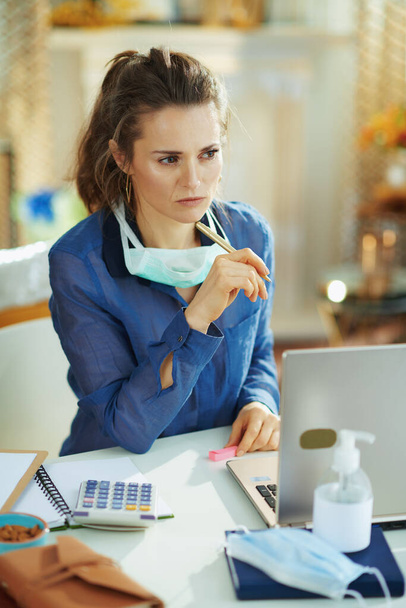 pensive 40 years old woman in blue blouse with medical mask working in temporary home office during the coronavirus epidemic in the modern house in sunny day. - Фото, изображение