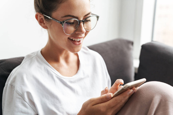 Image of young woman in eyeglasses smiling and typing on cellphone while sitting on sofa at living room - Фото, зображення