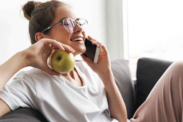 Image of excited nice woman in eyeglasses talking on cellphone and holding apple while sitting on sofa at living room - Φωτογραφία, εικόνα