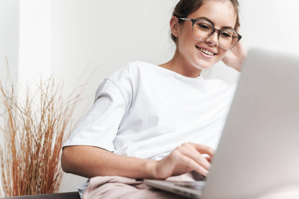 Image of smiling beautiful woman in eyeglasses using laptop while sitting on sofa at living room - Фото, зображення