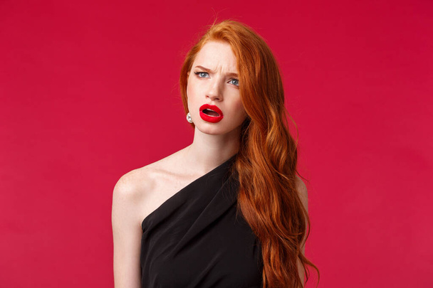 Close-up portrait of perplexed and confused young redhead woman seeing something strange, squinting and frowning as looking left troubled identify what is this, stand red background - Zdjęcie, obraz