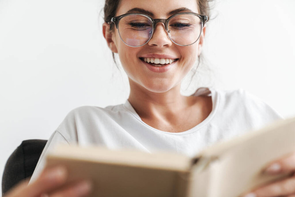 Image closeup of joyful caucasian woman in eyeglasses reading book while sitting on sofa at living room - 写真・画像