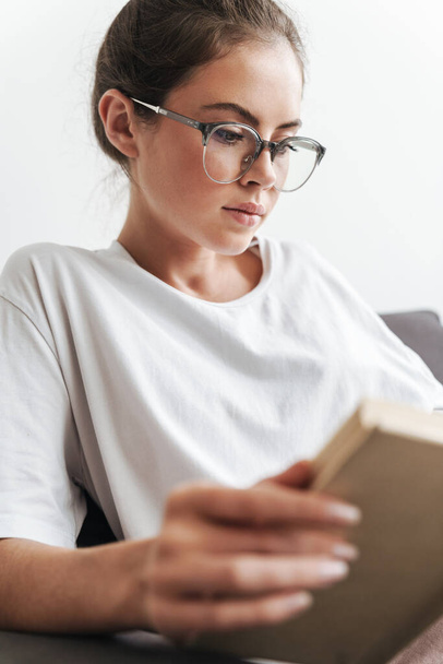 Image closeup of serious caucasian woman in eyeglasses reading book while sitting on sofa at living room - Foto, afbeelding