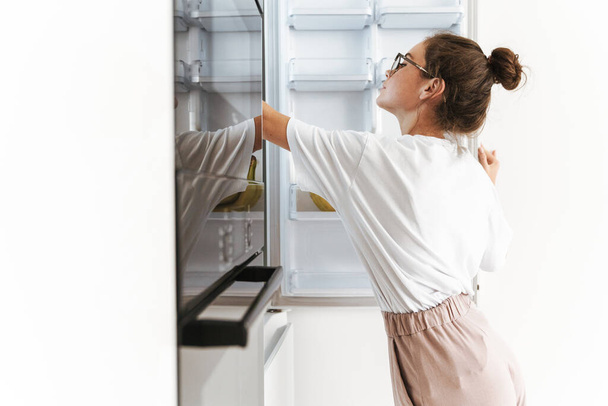 Image of beautiful young woman in eyeglasses looking at open fridge in sunny kitchen - Foto, imagen