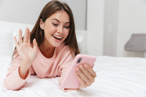 Cheerful lovely young girl wearing pajamas laying in bed at the bedroom, using mobile phone, having video chat - Photo, Image