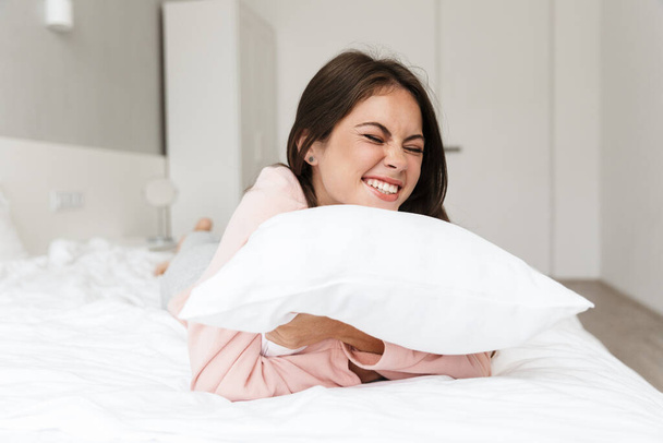 Cheerful lovely young girl wearing pajamas resting in bed at the bedroom, hugging pillow - Foto, imagen
