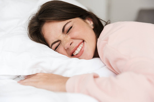 Cheerful lovely young girl wearing pajamas resting in bed at the bedroom, hugging pillow - Фото, зображення