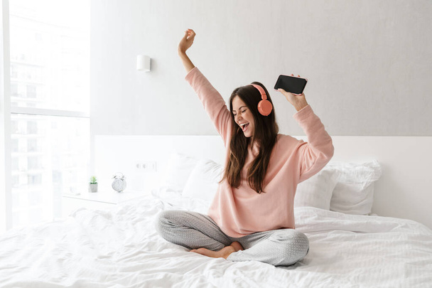 Cheerful lovely young girl wearing pajamas sitting on bed at the bedroom, listening to music with wireless headphones, holding mobile phone, dancing - Fotó, kép