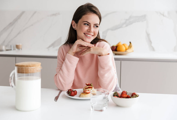 Beautiful smiling young girl having tasty healthy breakfast while sitting at the kitchen table - Фото, изображение