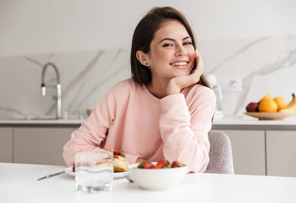 Beautiful smiling young girl having tasty healthy breakfast while sitting at the kitchen table - Fotoğraf, Görsel