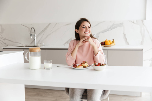 Beautiful smiling young girl having tasty healthy breakfast while sitting at the kitchen table - 写真・画像