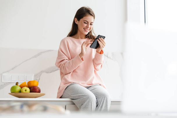 Cheerful lovely girl sitting on a kitchen desk with bowl of fruits, using mobile phone - Фото, зображення