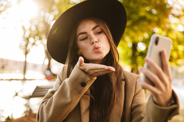 Portrait of gorgeous woman wearing coat and hat using cellphone while walking in autumn park - 写真・画像