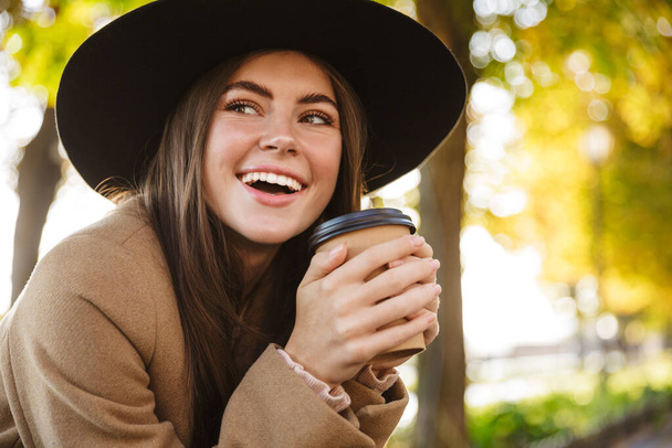 Portrait of happy woman wearing coat and hat drinking coffee while walking in autumn park - Foto, Bild