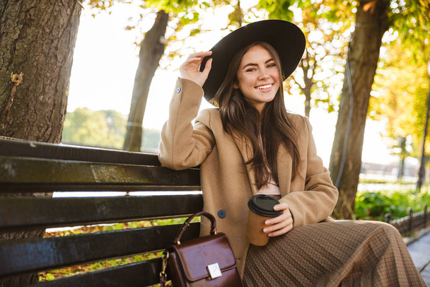 Portrait of tasteful woman wearing coat and hat sitting on bench with coffee cup in autumn park - Fotoğraf, Görsel