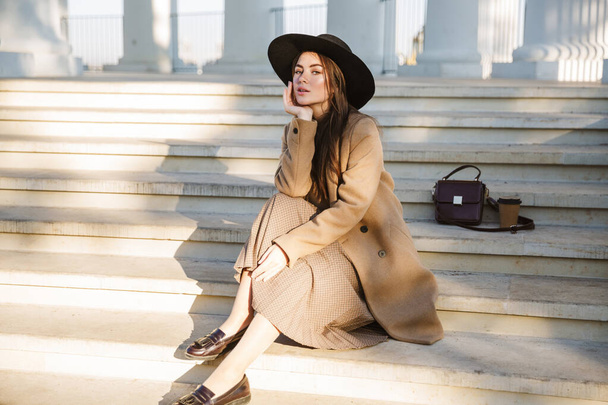 Portrait of adorable young brunette woman wearing coat and hat sitting on building stairs outdoors - Фото, зображення