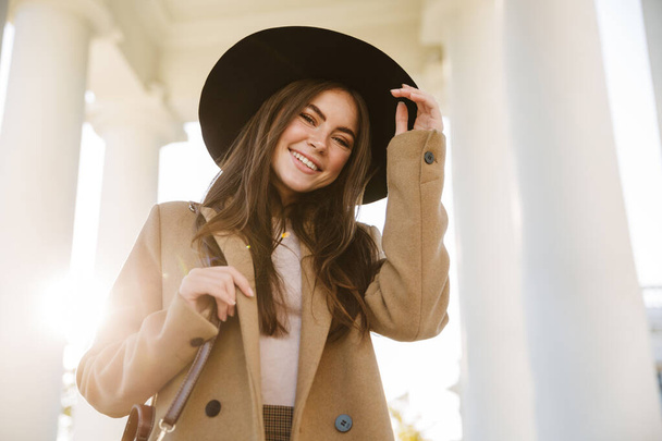 Portrait of adorable woman in autumn coat and hat smiling while standing by pillars outdoors - Fotografie, Obrázek