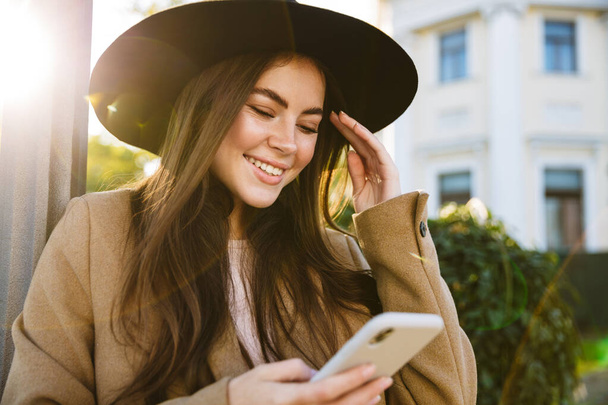 Portrait of gorgeous woman in autumn coat and hat holding smartphone while walking outdoors - Foto, Bild