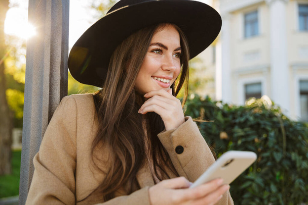 Portrait of pleased woman in autumn coat and hat holding smartphone while walking outdoors - Фото, зображення