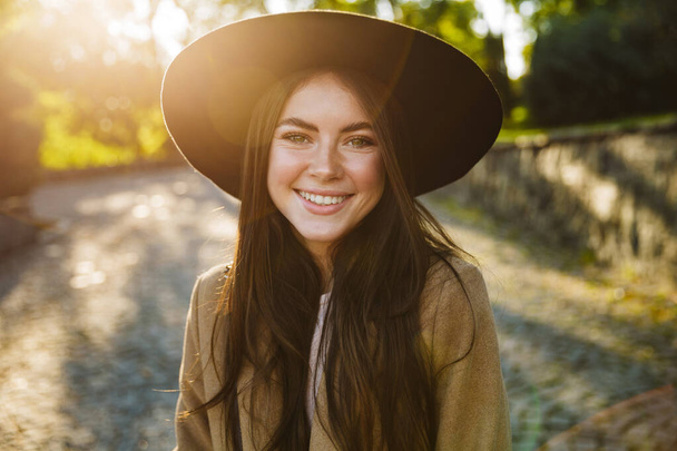 Image of satisfied woman in autumn coat and hat smiling while walking in green park outdoors - Foto, imagen