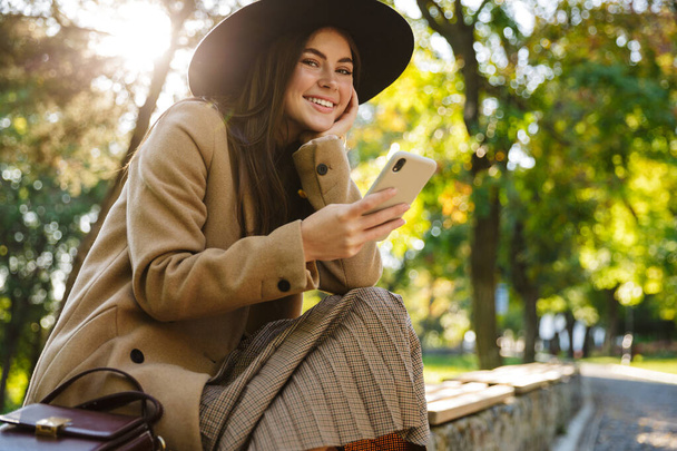 Image of positive woman in autumn coat and hat using smartphone while sitting on bench in park - Photo, image