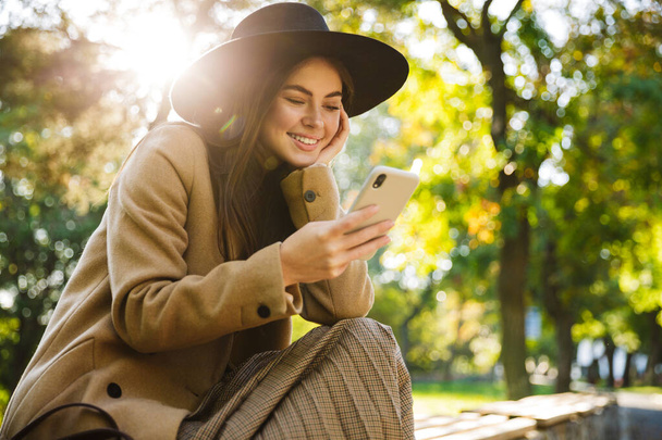 Image of joyful woman in autumn coat and hat using smartphone while sitting on bench in park - Zdjęcie, obraz