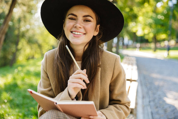 Image of caucasian woman in autumn coat and hat holding diary book while sitting on bench in park - Foto, Imagem