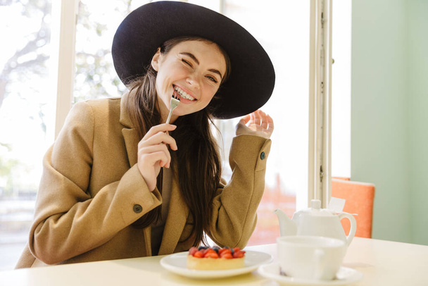 Image of charming young woman in autumn coat and hat eating sweet cake at indoor cafe - Foto, afbeelding