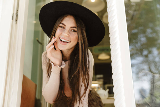 Image of brunette young woman in hat smiling while looking outside open window in cafe - Foto, imagen