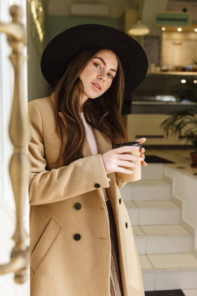 Afbeelding van verleidelijke jonge vrouw in herfst jas en hoed met koffiebeker in indoor cafe - Foto, afbeelding