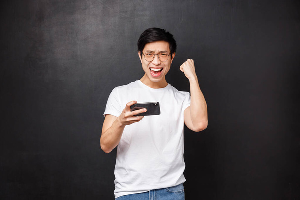 Technologie, gadgets a lidé koncept. Portrét triumfující a potěšené asijské chlap slaví vítězství nad přítelem v mmorpg online mobilní hra, pěst čerpadlo radostně, mistr smartphone arkády - Fotografie, Obrázek