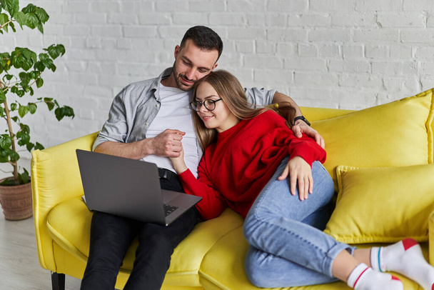 Joven linda pareja mirando a la pantalla del ordenador portátil sentado en el sofá amarillo
 - Foto, imagen