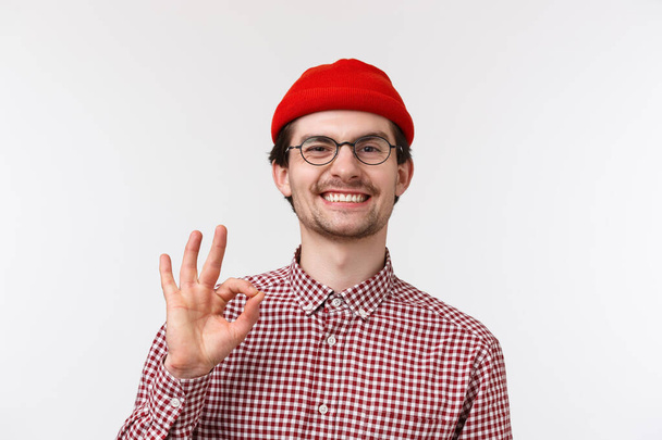 Close-up portrait of satisfied cool hipster guy in red beanie, glasses and checked shirt, smiling pleased, showing okay gesture, accept or agree with someones great idea, like plan, nod agreement - Foto, Imagem