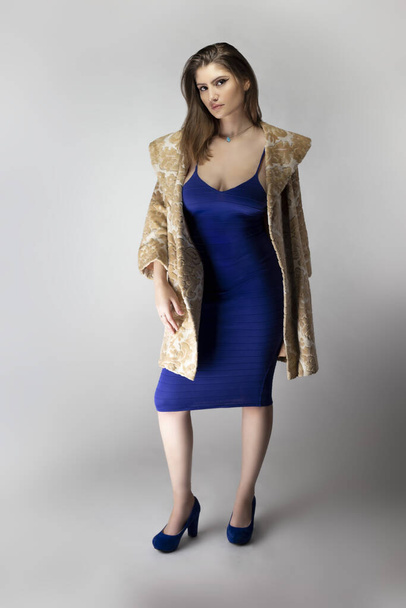 Female fashion model wearing a beige brocade wool or cotton coat and a blue sexy dress in studio catalog style.  Depicts spring or fall trends - Fotografie, Obrázek