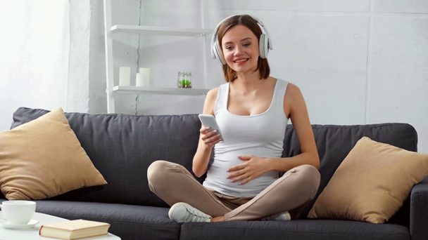 Vidám terhes lány fülhallgató gazdaság smartphone és énekel a kanapén  - Felvétel, videó