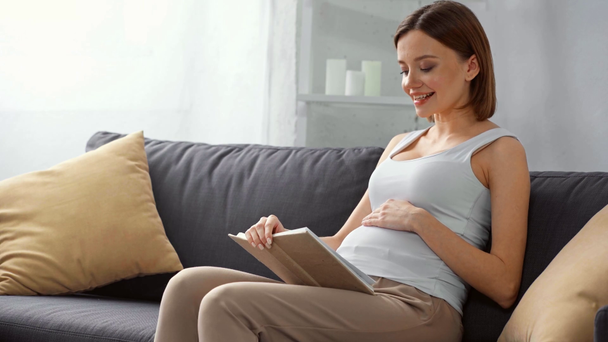 Usmívající se těhotná žena čtení knihy na gauči  - Záběry, video