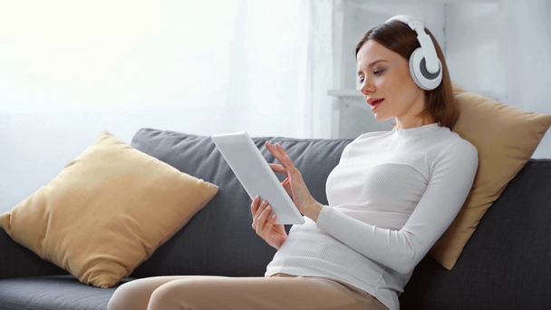 Kobieta w ciąży w słuchawkach za pomocą tabletu cyfrowego na kanapie  - Materiał filmowy, wideo