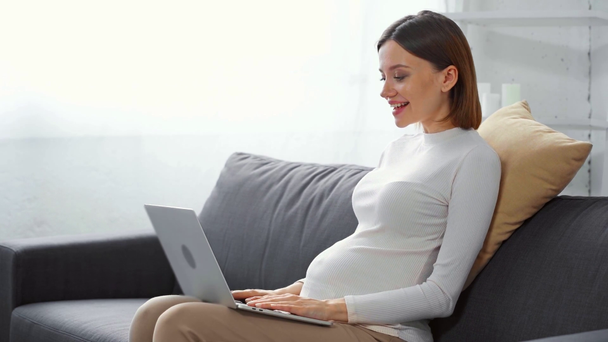 Vista lateral de la chica embarazada sonriente teniendo videollamada en el ordenador portátil en casa
 - Imágenes, Vídeo