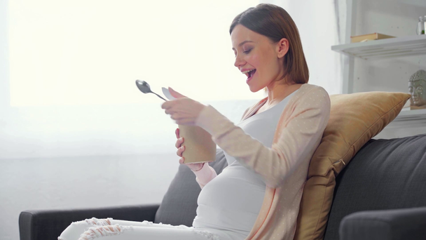 Πλευρική άποψη του χαρούμενα έγκυος γυναίκα τρώει παγωτό στον καναπέ  - Πλάνα, βίντεο
