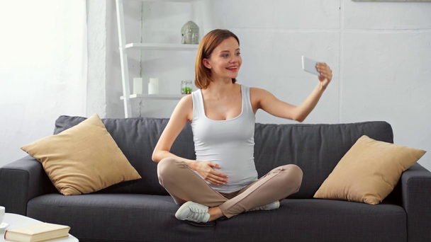 Счастливая беременная женщина делает селфи со смартфоном на диване
  - Кадры, видео