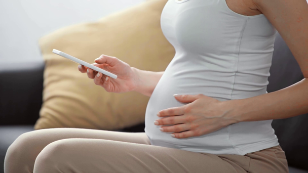 Καλλιεργημένη άποψη της εγκύου γυναίκας χρησιμοποιώντας smartphone στον καναπέ  - Πλάνα, βίντεο