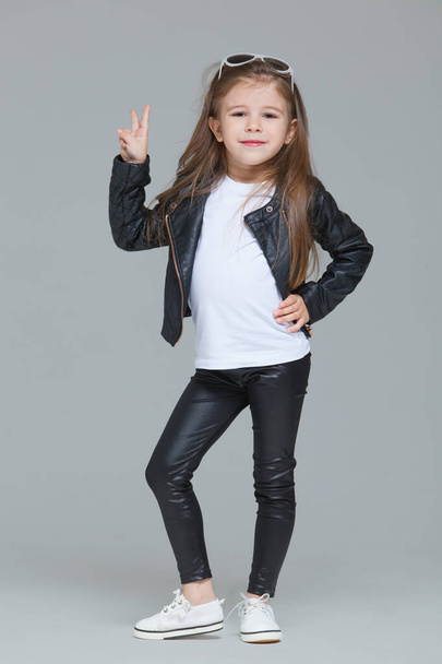 Baby girl with long hair in black leather jacket and leggings is posing in the studio in modern sunglasses on grey background - Foto, Imagem