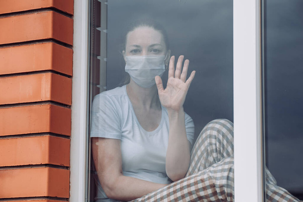 Home quarantine. A Caucasian woman is sitting at window in a medical mask , looking out, her hand on glass, wants to get out. protection against coronavirus infection, pandemics, outbreaks epidemics - Фото, изображение
