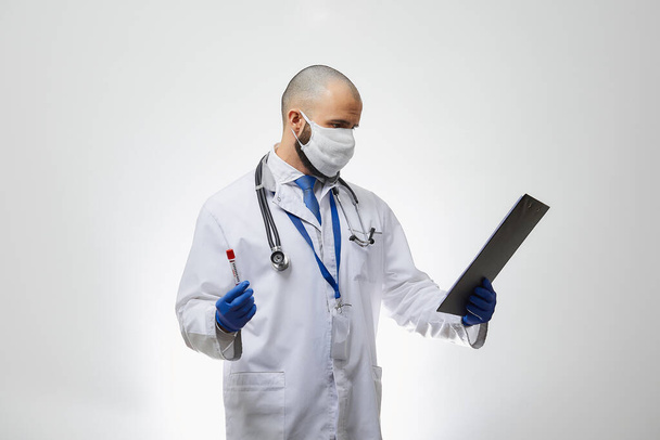 A doctor in a medical face mask reading information about a coronavirus blood test in his hand. A portrait of an infectious disease physician holding a positive COVID-19 blood sample and a clipboard. - Photo, Image